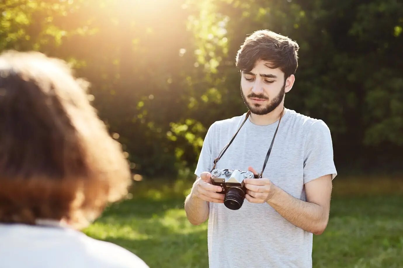Ein Mann mit Bart und stylischer Frisur fotografiert seine Freundin, die in der Natur posiert und sich die Bilder anschaut, die er mit seiner Retro-Kamera gemacht hat.